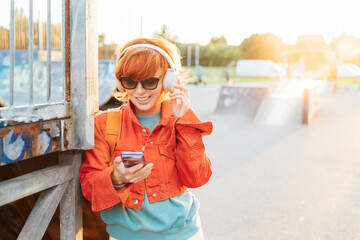 Smiling stylish woman in bright clothes, sunglasses wearing wireless headphones on her head and seting phone for listening music. Walk in city skate park on sunset. Hipster lifestyle. Selective focus.