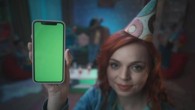 A girl in a festive hat shows a smartphone with a green screen, smiles, turns to the camera and nods her head in agreement. Close up of a girl with a smartphone on a blurred background of the room.