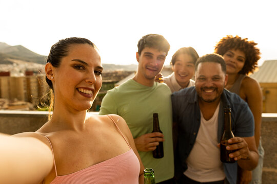 An interracial millennial group are taking a photo on a terrace while holding beers. The Caucasian girl is in the foreground picking up the smart phone.Concept of multiethnic group drinking alcohol.