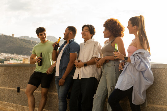 A group of 5 young interracial millennials are talking happily leaning against a wall of a one-story terrace while drinking beer. Concept of multiethnic group drinking alcohol, social drinkers.
