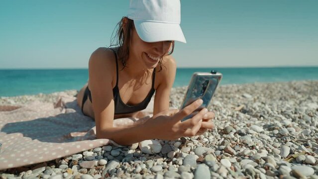 relaxed woman tourist in bikini sunbathes on sea beach close to water, unlocks her mobile phone and takes photo or video call. Blue Lagoon resort, Oludeniz, Turkey.