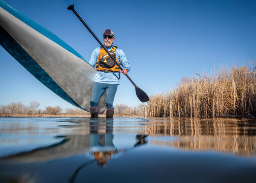 senior male paddler is launching a stand up paddleboard on a calm lake in early spring, frog perspective from an action camera at water level