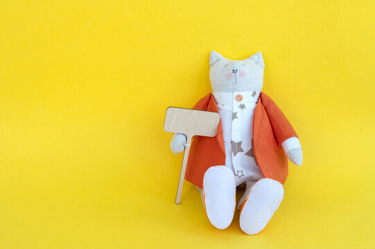 A soft toy cat in a jacket sits with a wooden sign with a place for text, on a yellow background