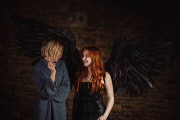 A girl with black angel wings and a woman with a cigarette laugh on a dark background.