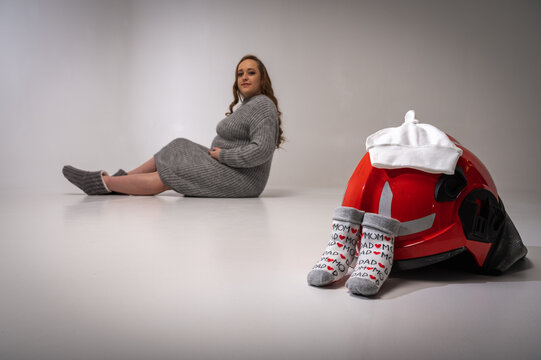 young pretty pregnant girl sits on a light background, isolated, fireman's helmet and small socks near her. Creating and waiting for a young firefighter family