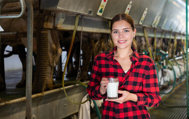 Confident young pretty female milker posing with glass of milk on dairy farm