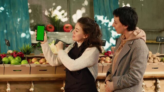 Two women looking at greenscreen display on smartphone, checking chroma key template on mobile phone app. Client and local farmer using isolated mockup copyspace template. Tripod shot.