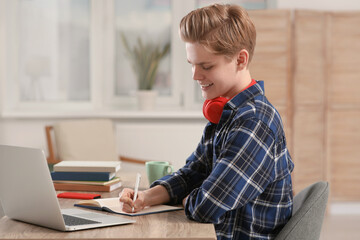 Online learning. Smiling teenage boy writing in notebook near laptop at home