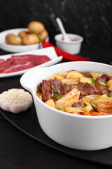 Pot of delicious vegetable soup with meat, noodles and ingredients on black wooden table, closeup