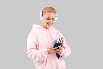 Fototapeta premium Young woman in headphones with books using mobile phone on grey background