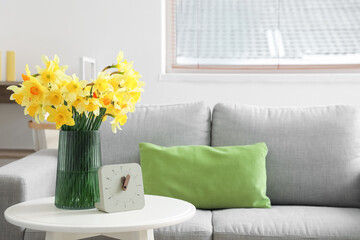 Vase with blooming narcissus flowers and clock on coffee table in interior of living room