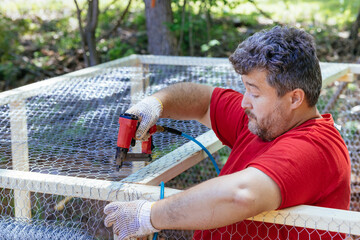 An individual builds wooden domestic chicken coop on farm by nailing metal mesh to frame building montage afterward © ungvar