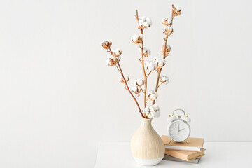 Cotton sprigs in vase with books and alarm clock on table near white wall