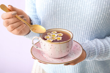 Beautiful young woman with cup of chamomile tea near lilac wall