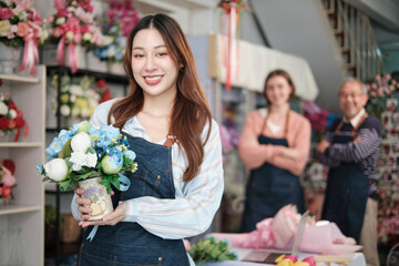 Fototapeta premium Beautiful young Asian female florist worker with vase of blossom smiles and looks at camera in front of colleagues team in colorful flower shop, small business occupation, happy SME entrepreneur.