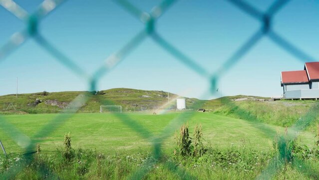 View Through Steel Fence Of Sprinkler Spraying Water On Grass In Football Field In Lovund, Norway. wide