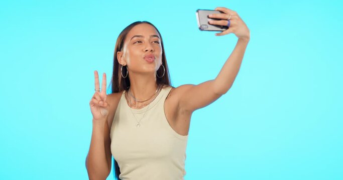 Woman, selfie and peace sign with kiss face, influencer and social media post with memory on blue background. Mockup space, energy with young female person happy and wink in picture with live stream