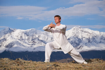 Wushu master in a white sports uniform training kungfu in nature on background of snowy mountains.