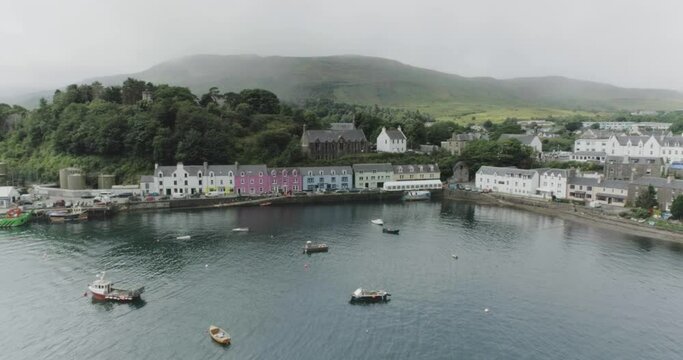 Wide drone shot showing moored boats, colourful houses and misty mountains in a bay on Loch Portree on the Isle of Skye