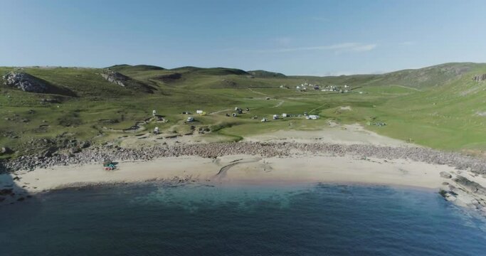 Slow wide drone shot of a perfect white sandy beach with clear blue waters and wild camping vehicles in the distance along the Scotland North Coast 500 Route