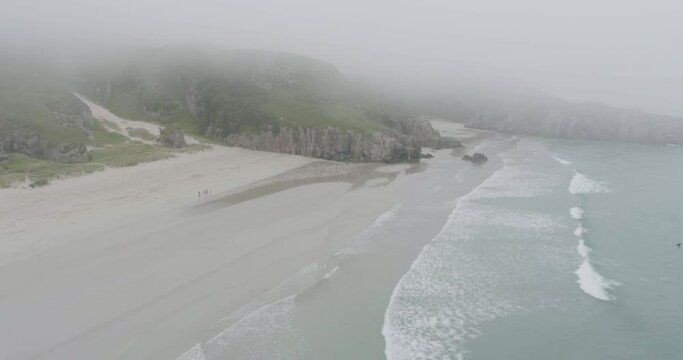 Slow drone reveal shot of surfers, big waves and misty weather on Ceannabeinne Beach on the Scotland North Coast 500 Route which is the North Sea