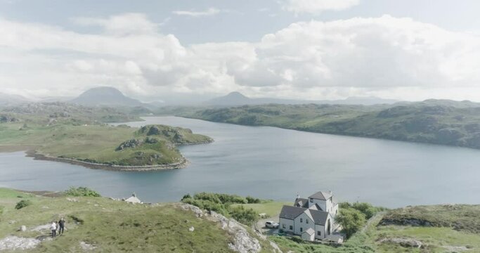 Slow drone panning shot of a house situated on a hill alongside a loch in scotland and tall mountains and clouds in the distance and sun shining
