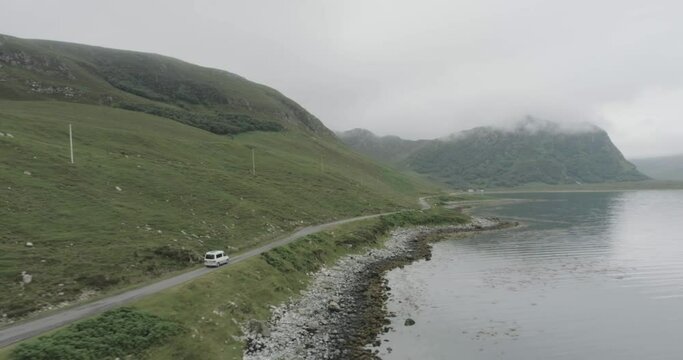Drone tracking shot of a campervan on a single track road during a trip of the Scotland North Coast 500 with a Loch and cloudy topped mountains in view