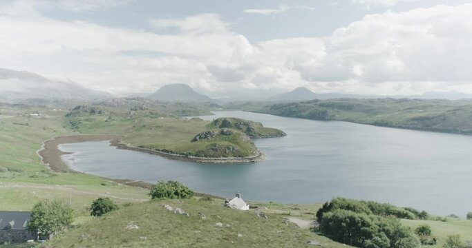 Slow drone panning shot of a Loch in Scotland with houses in the foreground, tall mountains and clouds in the distance and sun shining