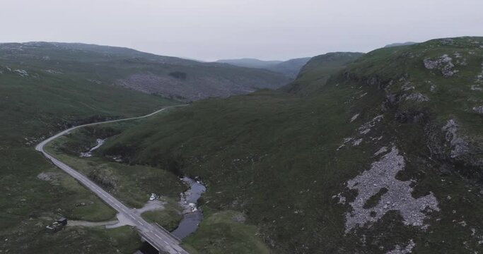Wide drone shot of a campervan parked up along a river on a remote road surrounded by hills at dusk in scotland