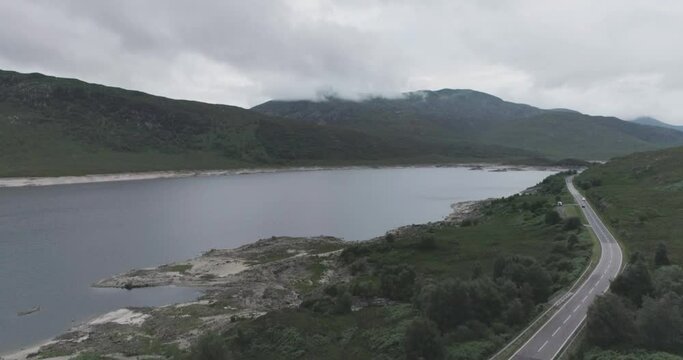 Super wide pull back drone shot of Loch Beag showing a campervan wild camping, BBQ smoke, coastal road and cloudy mountains in the background in the Scottish Highlands