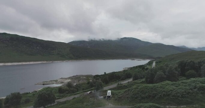 Wide drone shot showing Loch Beag and a campervan parked up for the night and a BBQ on the Loch edge with cloudy mountains in the background