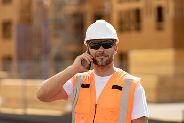 Builder call with phone with hardhat helmet on construction site. Construction builder worker in builders uniform near construction building.