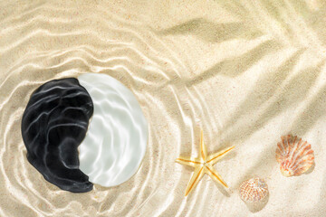 sun lights with hard shadow of palm leaf in wavy water on abstract sand background with shell, starfish and sign Yin-Yang stones, beautiful abstract spa concept