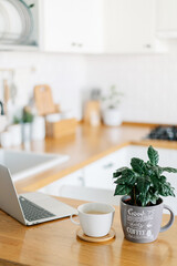 Cup of cofee and laptop on wooden table, view on white kitchen in modern style