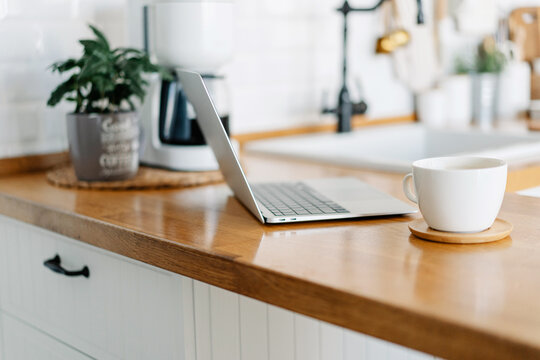 Cup of cofee and laptop on wooden table, view on white kitchen in modern style