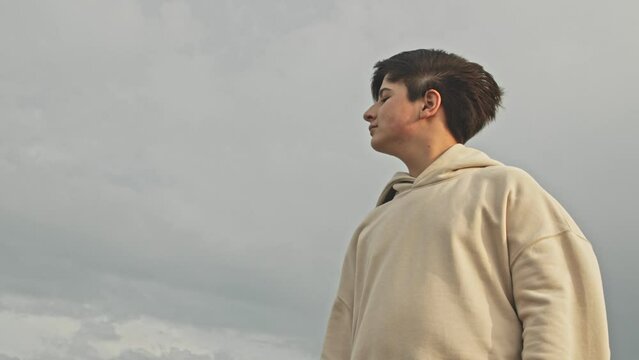 Slow motion Stylish teenager boy stands in the strong wind in the rays of the sunset. His clothes flutter against the sky
