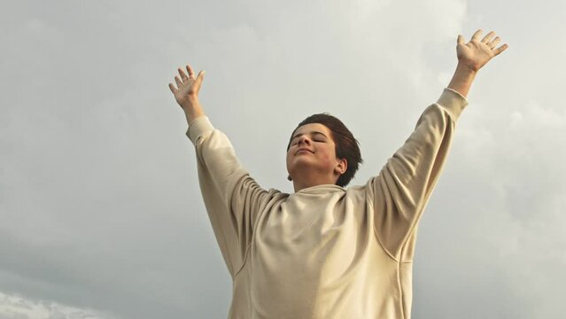 Wind catcher. Slow motion Stylish teenager boy stands in the strong wind with his hands up and smiles in the rays of the sunset. His clothes flutter against the sky