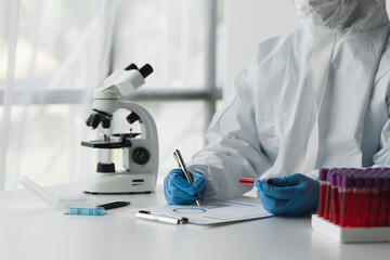 A scientist or microbiologist wearing PPE and a face mask holds a test tube with blood from a...