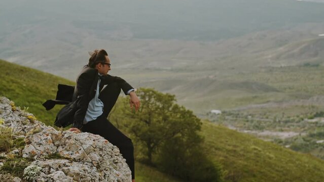 An adult man in a business suit fluttering in the wind sits on a ledge of a hill against the backdrop of a mountain range. Dreamer. Traveler