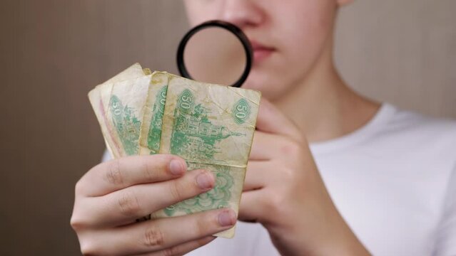 Teenager Examining Old Ukrainian Money, Coupons through a Magnifying Glass. Close up. Serious curious boy looks at signs, symbols, numbers on banknotes. Hobby. Grey blurred background. Collecting.