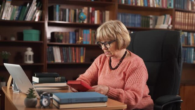 An elderly woman is sitting at her workstation, talking to someone via webcam. She holds a red notebook in her hands, and she seems to be sharing something interesting about it.