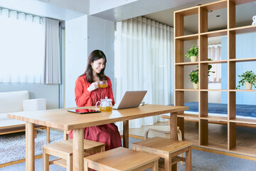 自宅のデザイナーズ住宅のリビングのテーブルでノートパソコンを見ながら笑顔で紅茶を飲む30代の女性