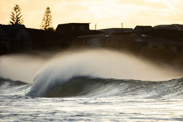 huge stormy shore break wave crashing on a beach at sunset