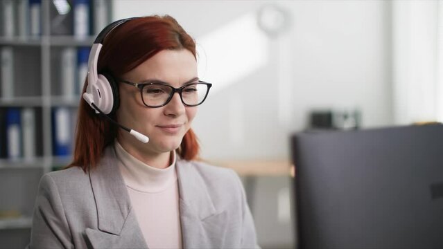 customer service, attractive woman in glasses works in a call center and communicates with a client using a headset while sitting at a computer in office