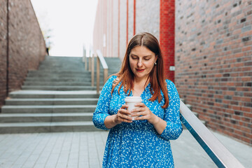 Cheerful young woman wearing blue dress walking outdoors, happy girl holding takeaway coffee cup on city background. Urban lifestyle concept.