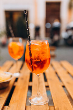 Aperol Spritz Cocktail. Alcoholic beverage based on table with ice cubes and oranges outdoors. Served cocktail with orange slice and straw placed on wooden table of sidewalk cafe in Italy