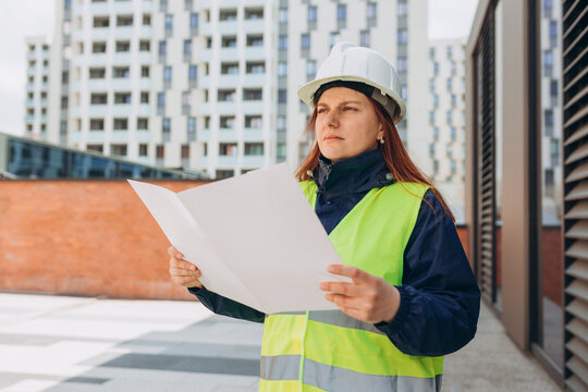 Architect with paper folder with documents at a construction site. Woman constructor wearing helmet and safety yellow vest. Women are planning new building project. Modern exterior