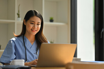 Smiling female doctor reading electronic medical records on laptop computer while sitting at modern workplace