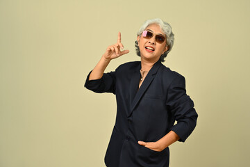 Cheerful senior grey haired businesswoman standing over beige background pointing finger up