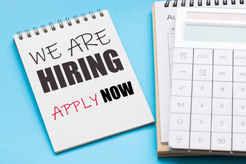 WE ARE HIRING APPLY NOW words in office notebook next to calendar and calculator.